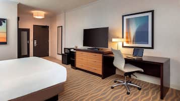 Una habitación de hotel con cama, escritorio con computadora, televisor, silla, lámpara y un cuadro en la pared.