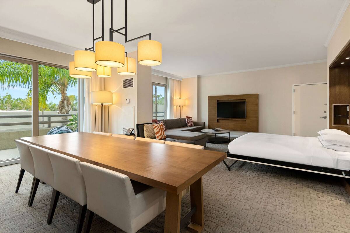 Una habitación de hotel moderna con una mesa de comedor de madera, un sofá, un televisor montado en la pared y una cama.
