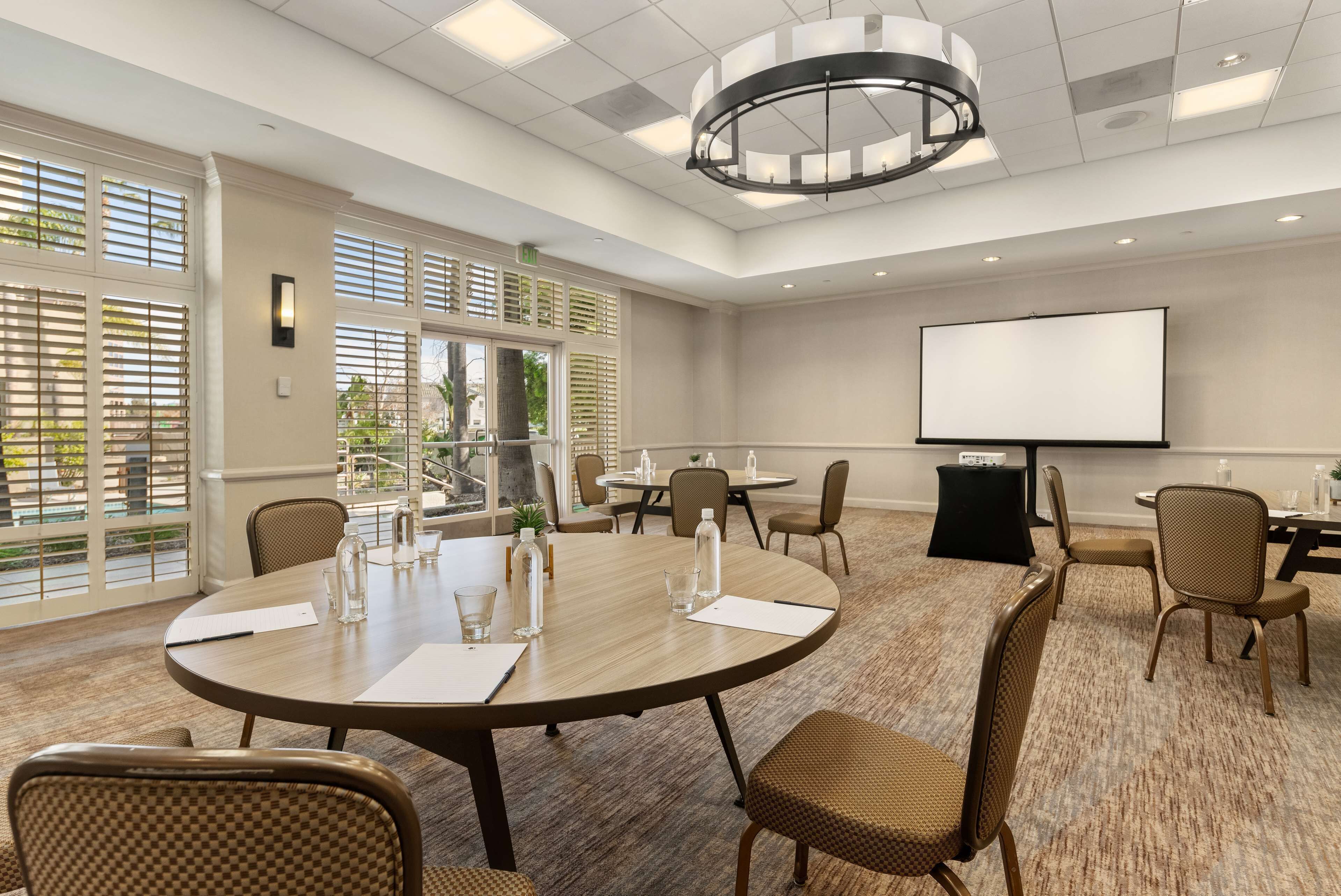 A conference room with a round table, chairs, a projector screen, and a large window with blinds.
