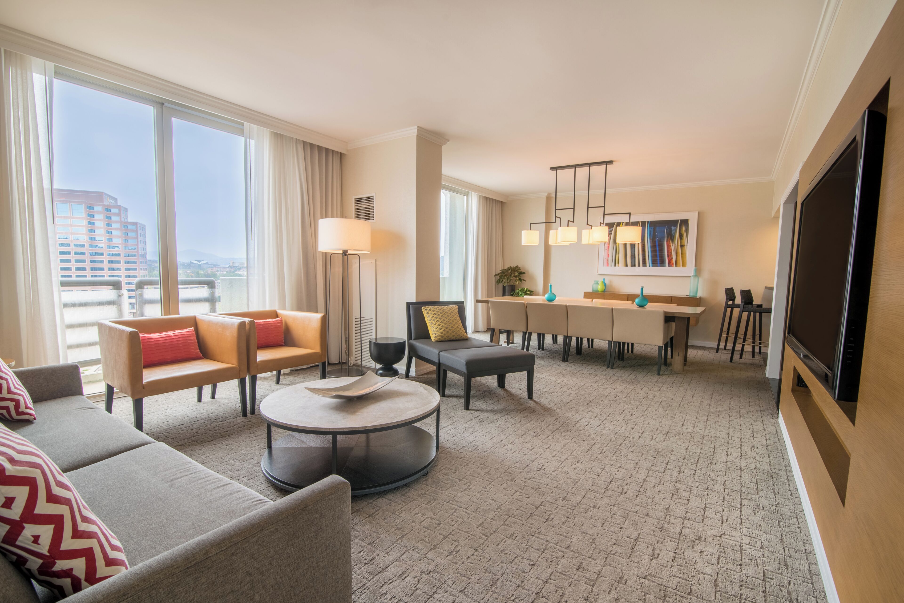 A hotel room with a sofa, armchair, round coffee table, dining area, and a TV.