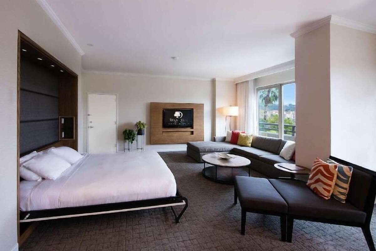 Habitación de hotel con cama, sofá, una mesita redonda, una silla, televisor montado en la pared y ventana con vistas.