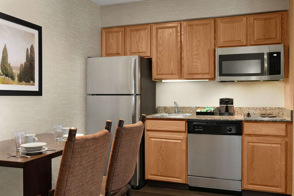 A modern kitchen with wooden cabinets, stainless steel appliances, and a dining area with chairs and a table set for two.