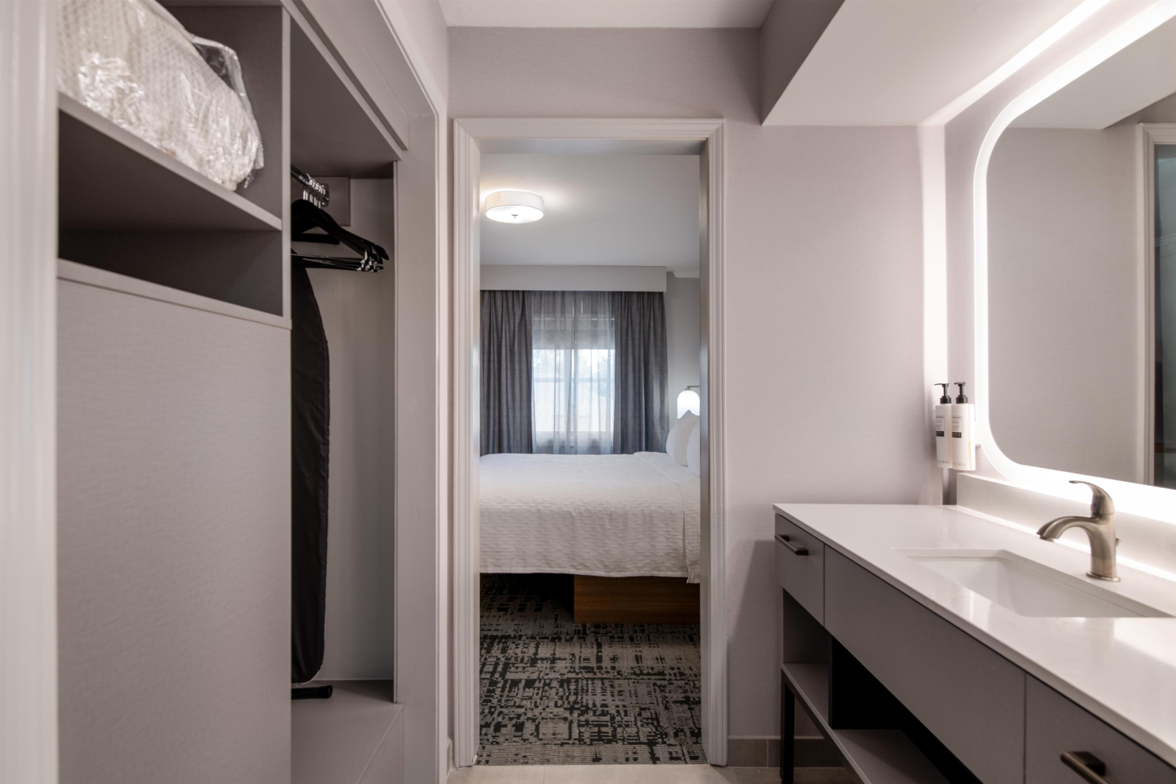 A hotel room with a walk-in closet, a bed with a white duvet, a bathroom with a modern sink and mirror, and a patterned rug on the floor.