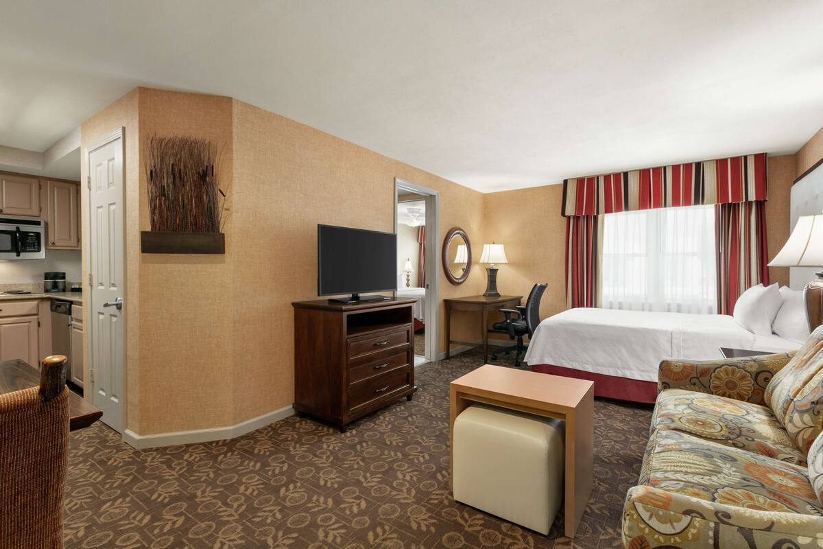 A hotel room with a bed, a desk, a television, a chair, a sofa, a small table, and a microwave in the kitchen area.