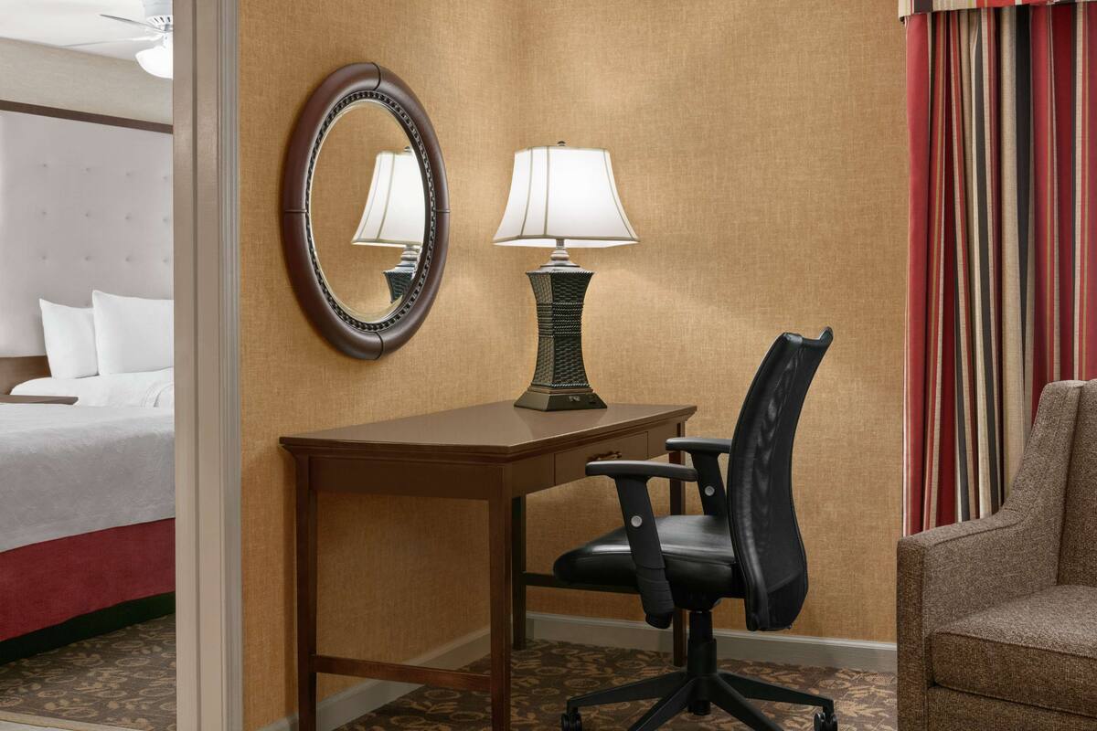 A hotel room with a bed, a desk with a lamp, a chair, a mirror, and a window with curtains.