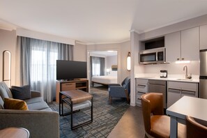 A hotel room with a TV, sofa, armchair, and a kitchenette with a microwave and coffee maker.