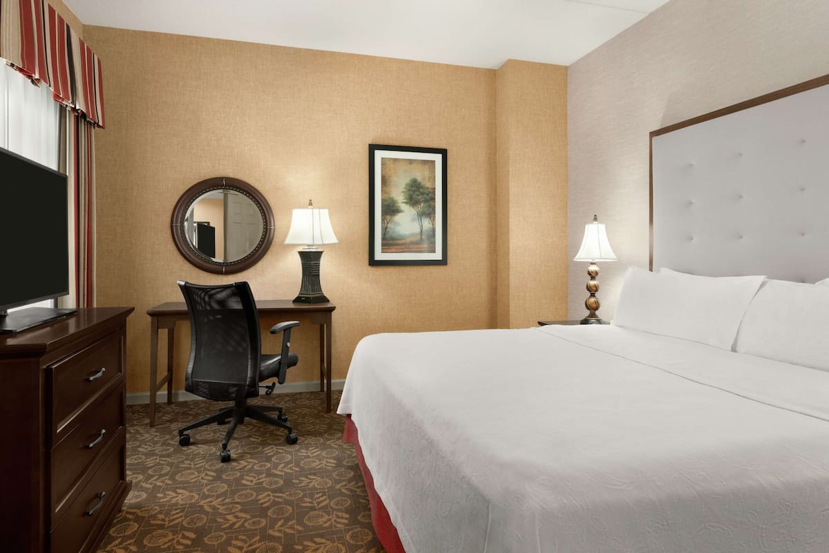 A hotel room with a bed, a desk with a chair, a television, a lamp, a framed picture on the wall, and a dresser.