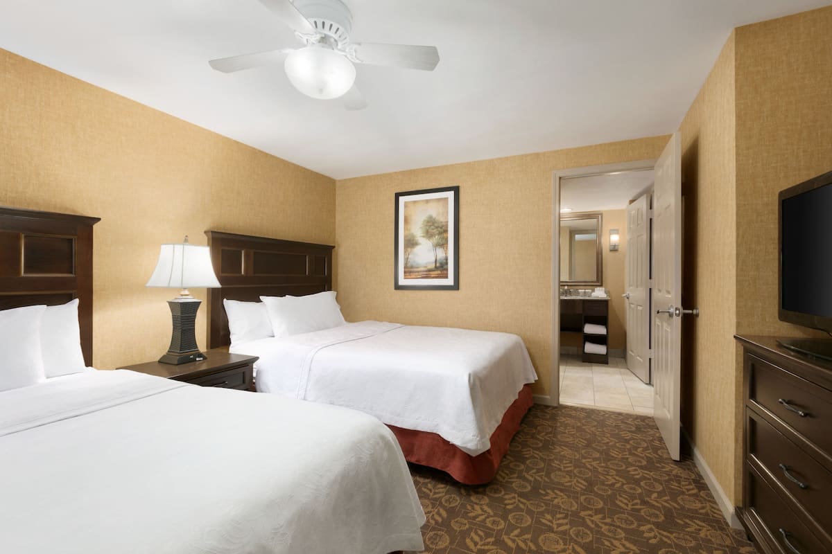 A hotel room with two beds, a ceiling fan, a painting, a bathroom, and a TV.