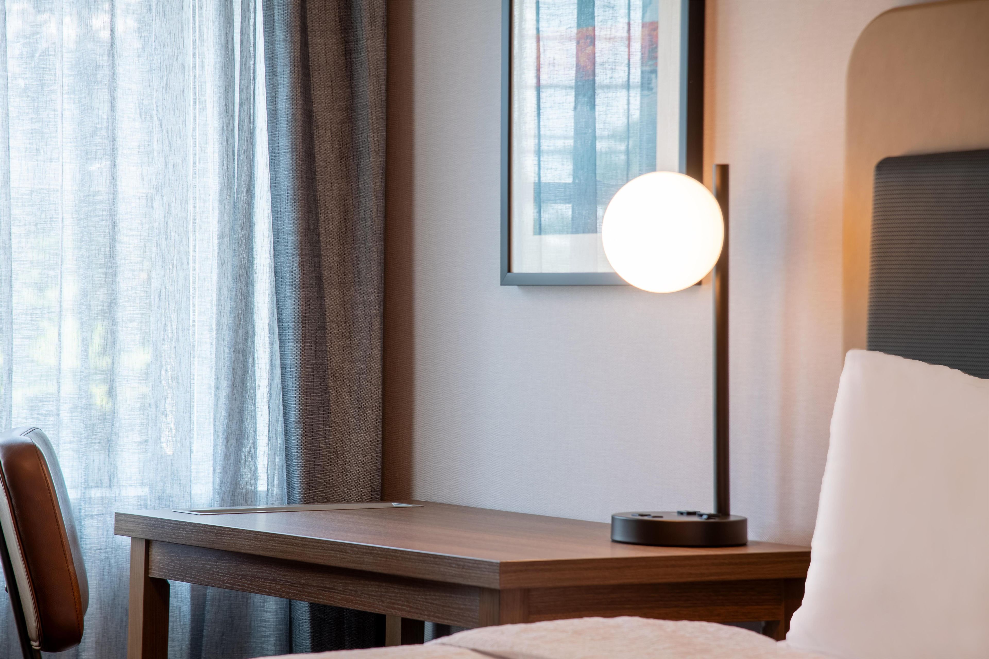 A hotel room with a bed, a desk, a chair, a lamp, and a window with curtains.