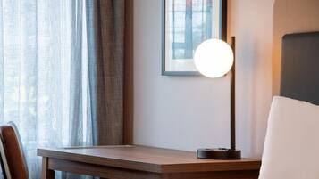 A hotel room with a bed, a desk, a chair, a lamp, and a window with curtains.