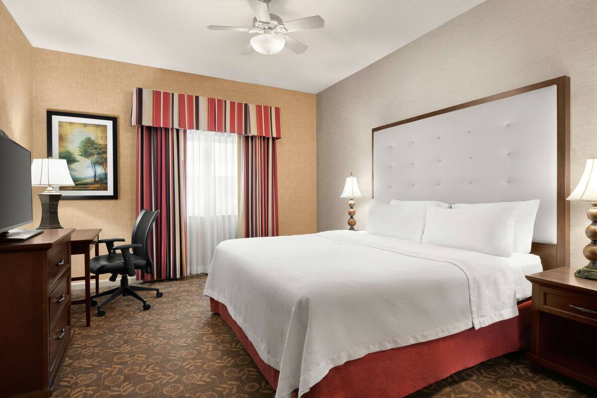 A hotel room with a large bed, a desk with a chair, a television, and a ceiling fan.