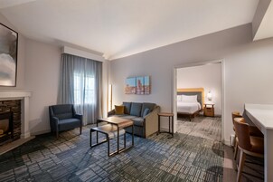 Habitación de hotel con sofá, sillón, mesita pequeña y cama con mesita de noche.