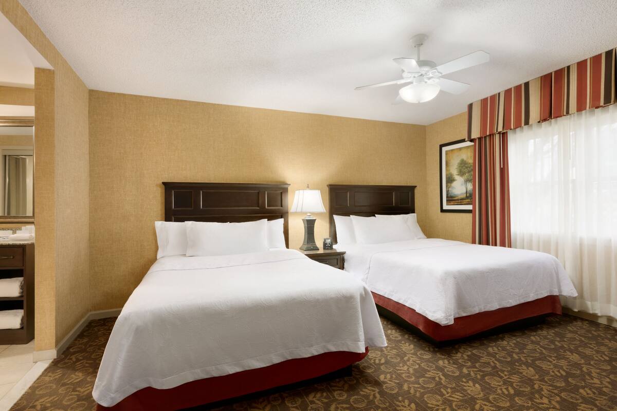 A hotel room with two beds, a ceiling fan, a nightstand with a lamp, and a window with curtains.