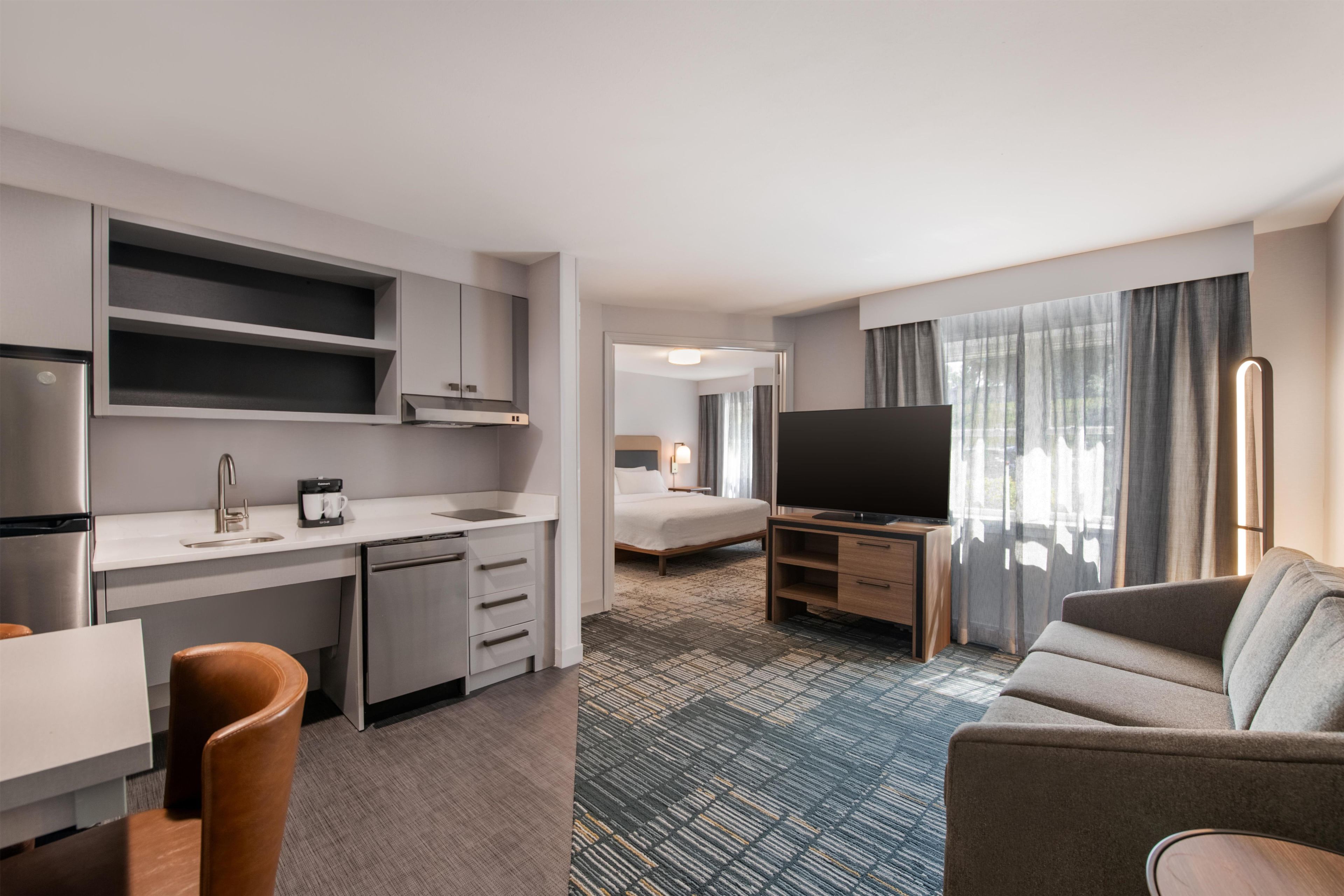 A modern hotel room with a kitchenette, a flat-screen TV, a sofa, and a bed.