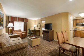 A hotel room with a living area, a dining table, a TV, a kitchen area, and a large window with curtains.