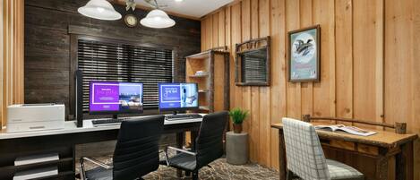 A modern office with two desks, black office chairs, a wooden desk with a plaid chair, a potted plant, and a poster on the wall.