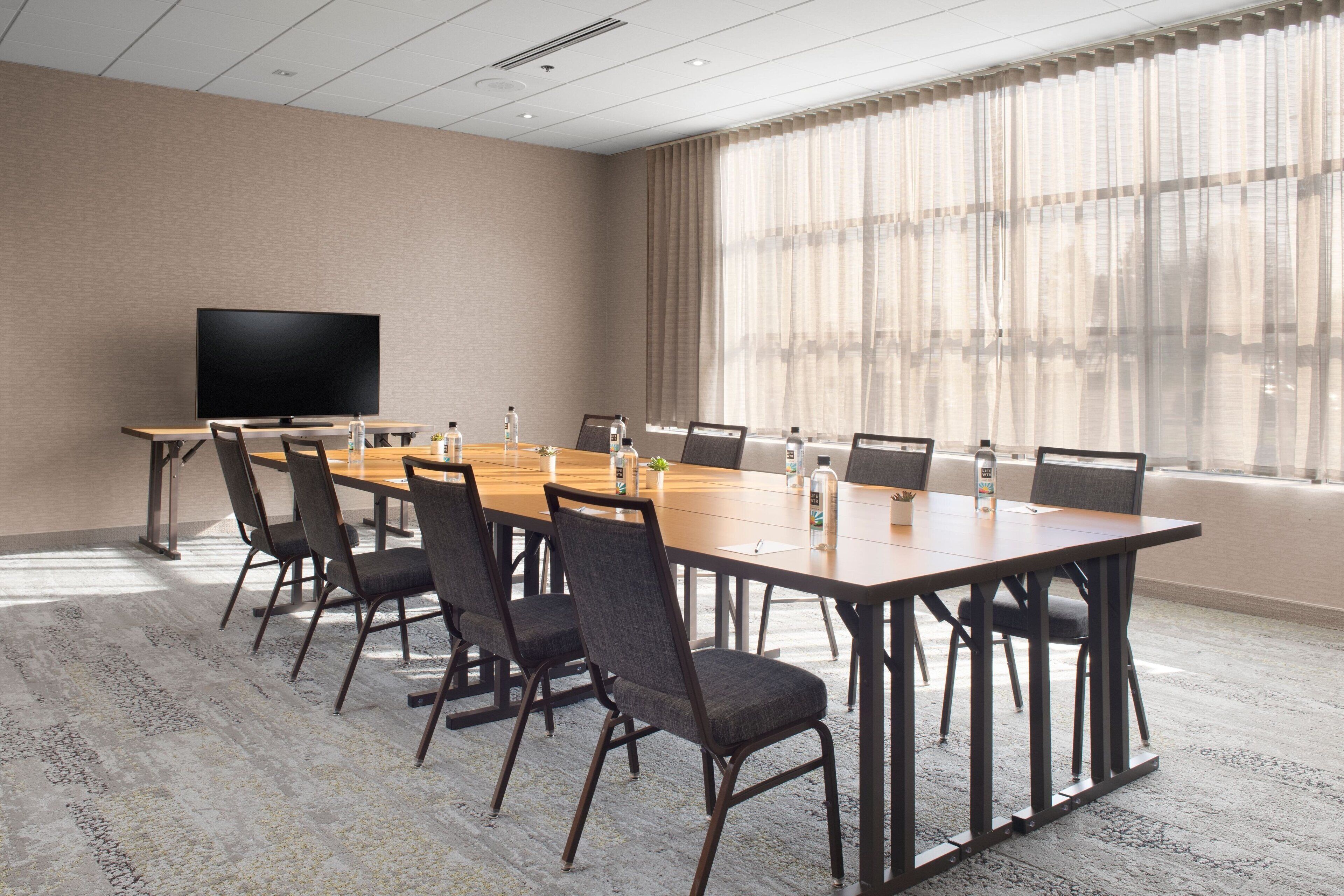A conference room with a long table, chairs, a flat-screen TV, and a large window with sheer curtains.