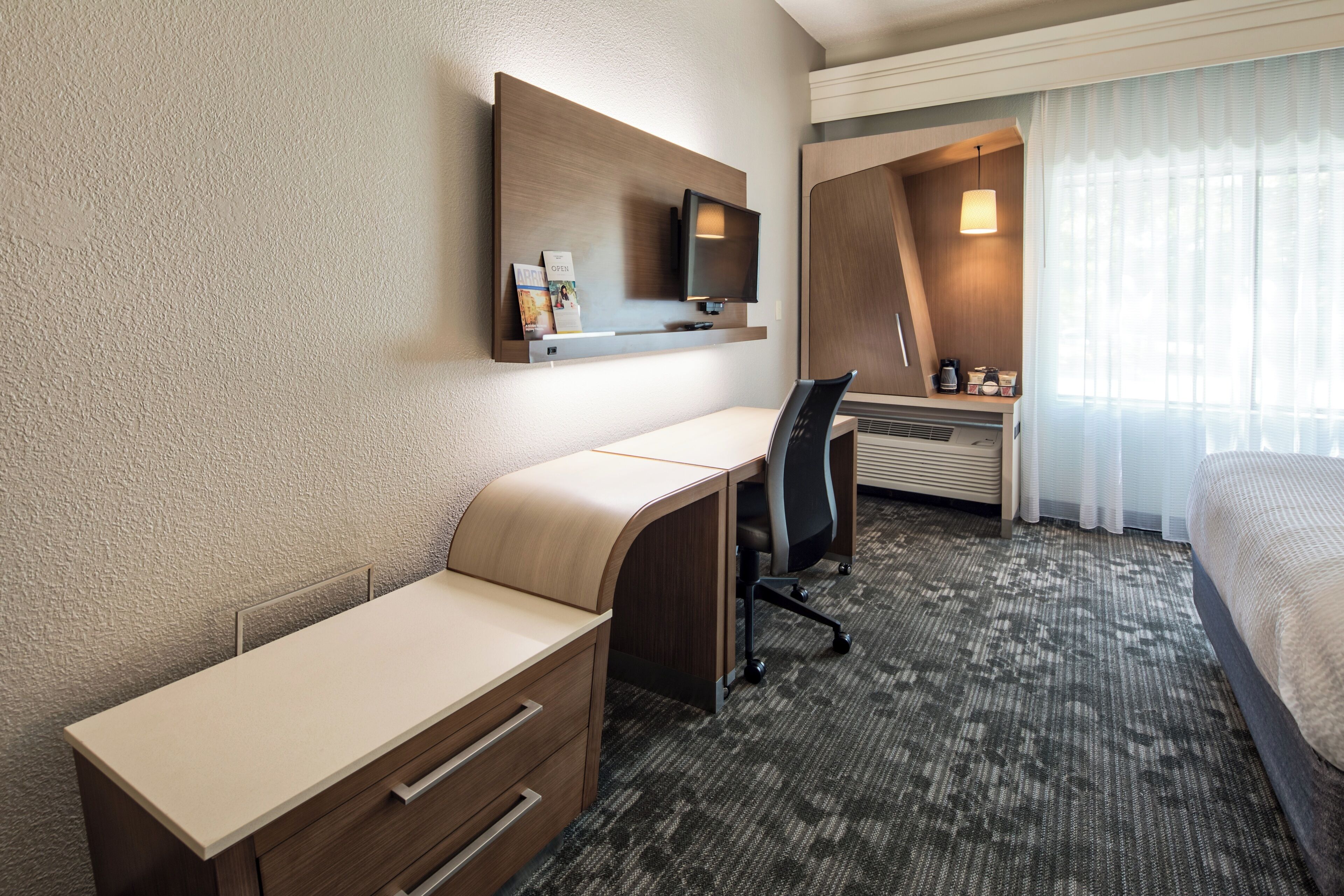 A hotel room with a bed, a desk with a chair, a television, and a window with curtains.