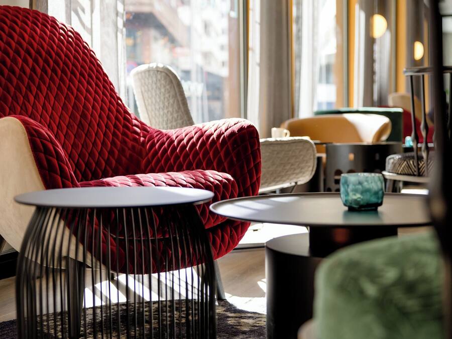 Un área de estar moderna con una butaca tapizada de rojo, una silla blanca y una silla verde. Hay dos mesitas redondas con patas negras.