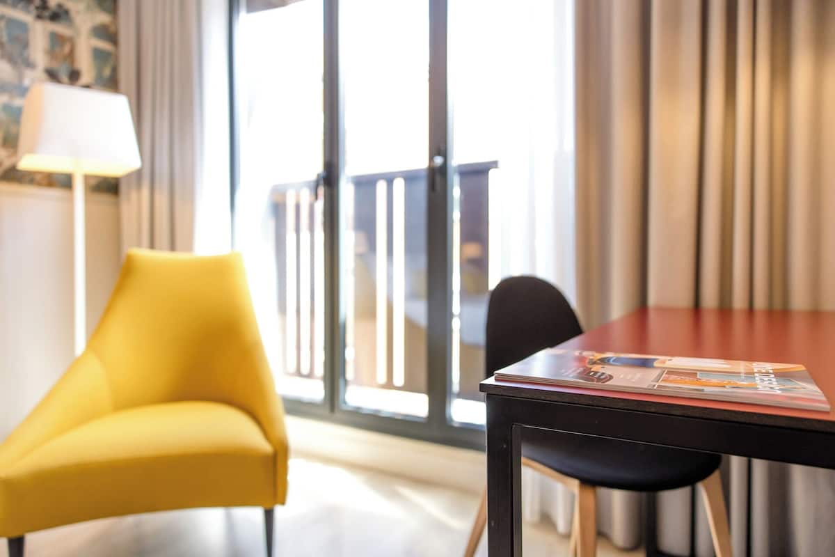 A hotel room with a yellow armchair, a wooden table with a magazine, and a view of a balcony through large windows.