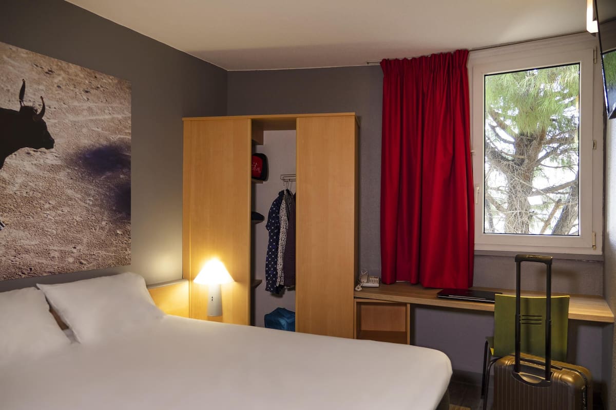 A hotel room with a bed, a desk, a wardrobe, a suitcase, a lamp, and a window with a view of trees.