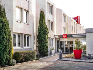 Entrada de hotel con una bandera roja, fachada de edificio moderno y un jardín bien cuidado.