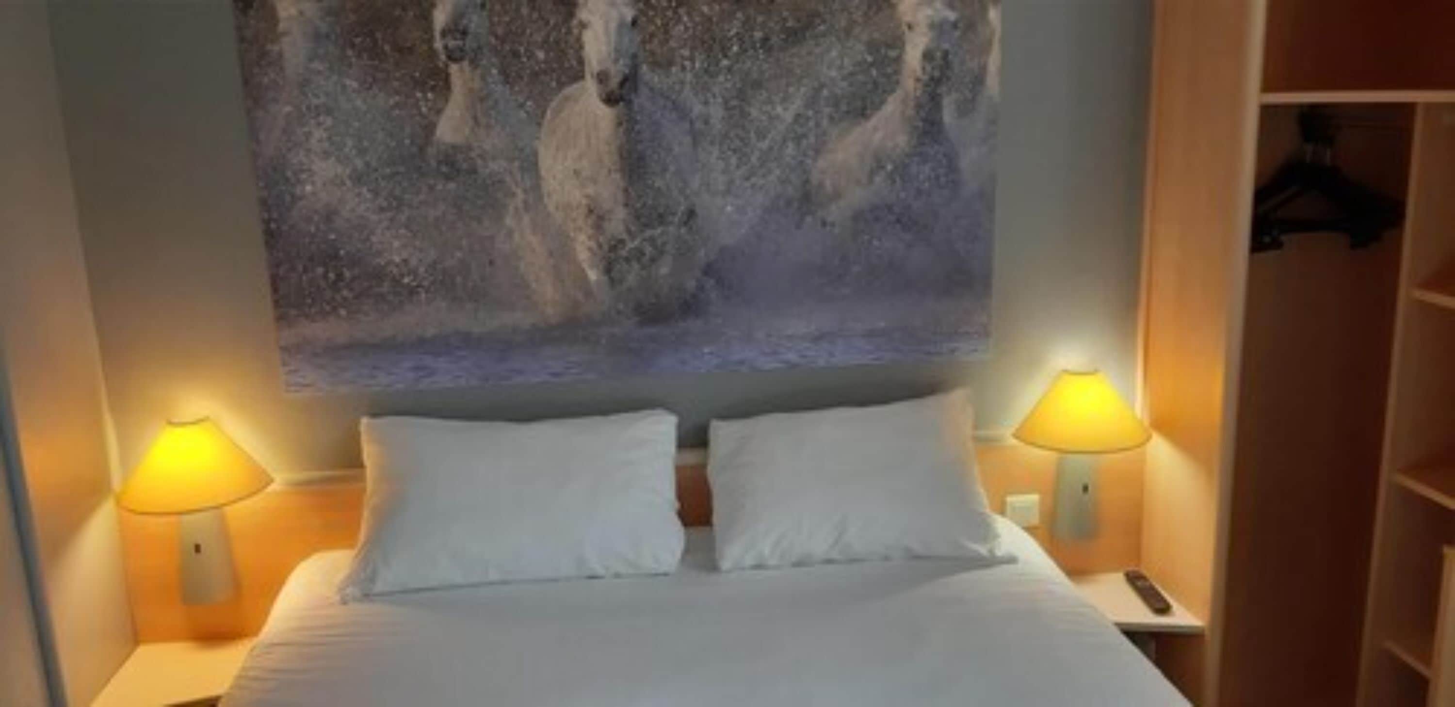 A hotel room with a bed, two bedside lamps, a painting above the bed, and a wardrobe to the right.