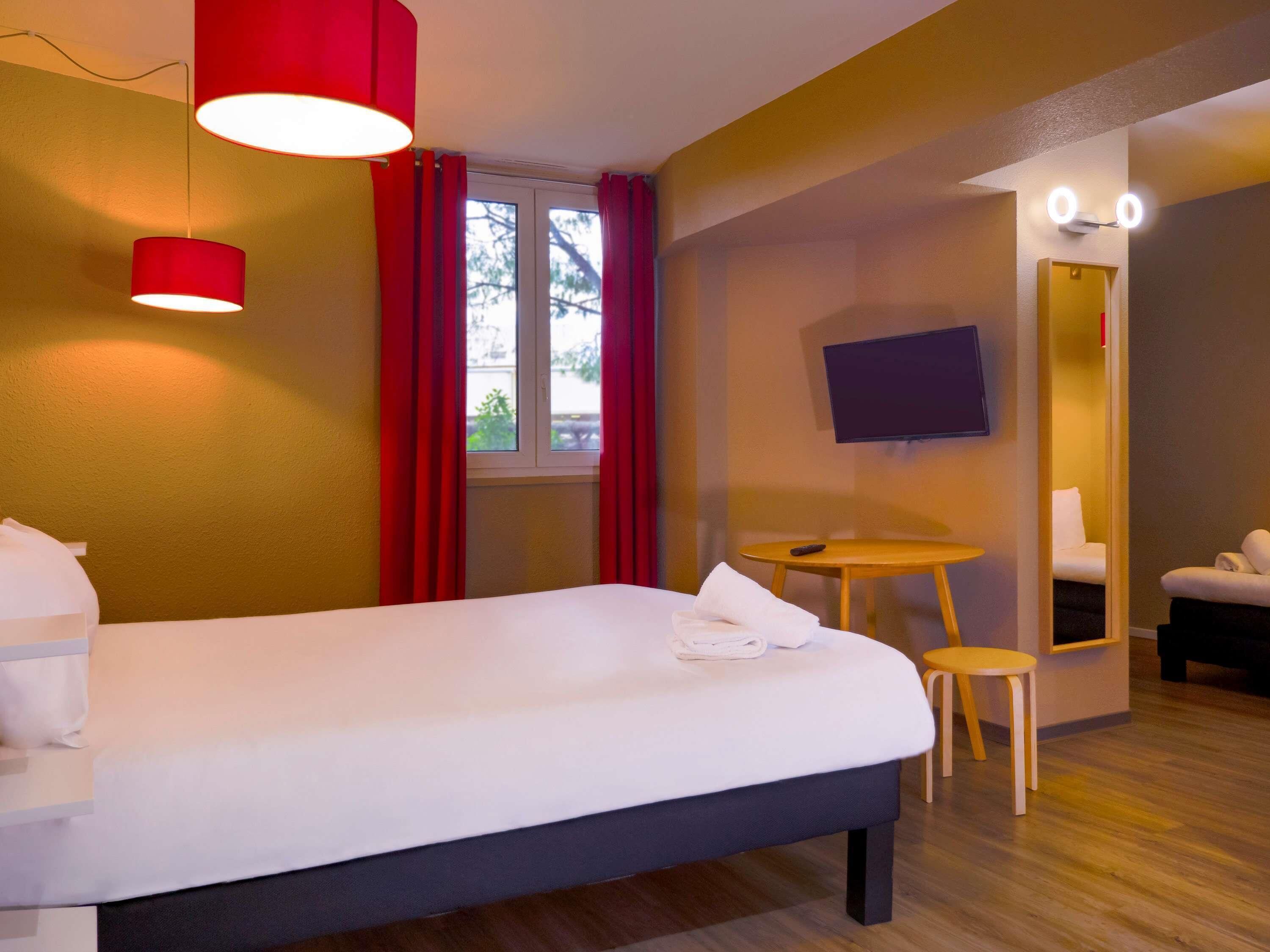 A hotel room with a bed, a TV, a small table with a chair, and a window with curtains.