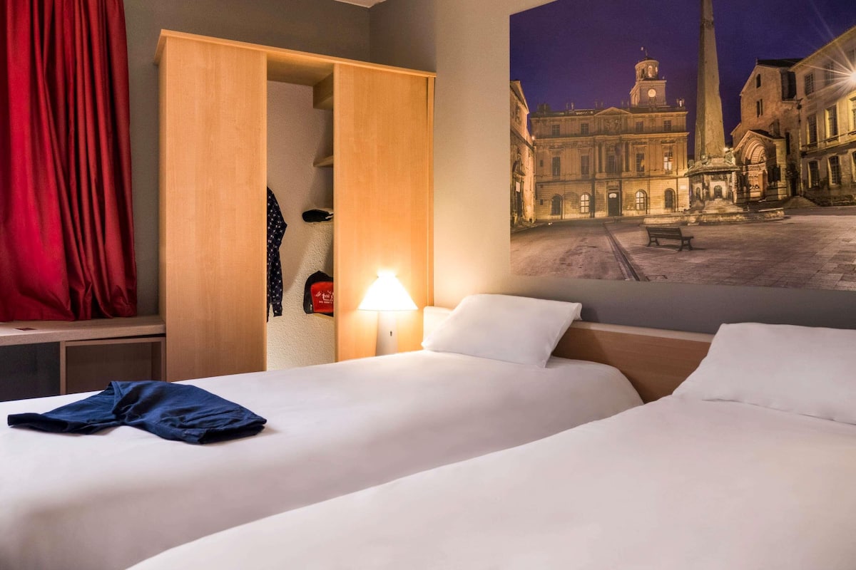 A hotel room with a bed, a nightstand, a lamp, a wardrobe, and a wall mural of a cityscape.