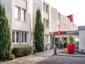 Entrada de hotel com uma bandeira vermelha, fachada de um edifício moderno e área de jardim bem cuidada.