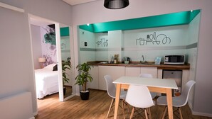 Ein kleines Hotelzimmer mit Küchenzeile, einem Essbereich mit Tisch und Stühlen, einem Bett und einer Zimmerpflanze.