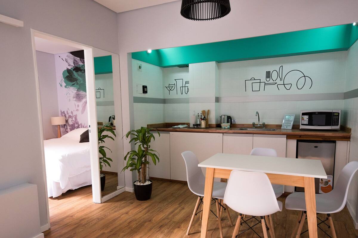 A compact hotel room with a kitchenette, a dining area with a table and chairs, a bed, and a potted plant.