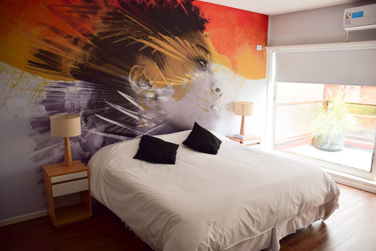 A bedroom with a large bed, two bedside tables, a wall mural, and a window with a view.