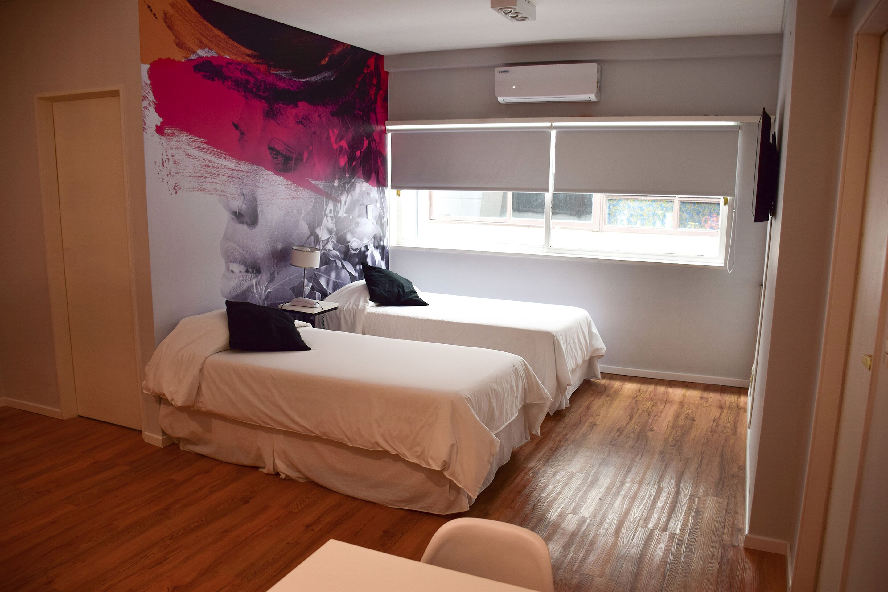Habitación de hotel con dos camas, un cuadro abstracto grande en la pared, una ventana con persianas y suelo de madera.
