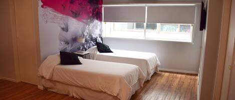 Habitación de hotel con dos camas, un cuadro abstracto grande en la pared, una ventana con persianas y suelo de madera.