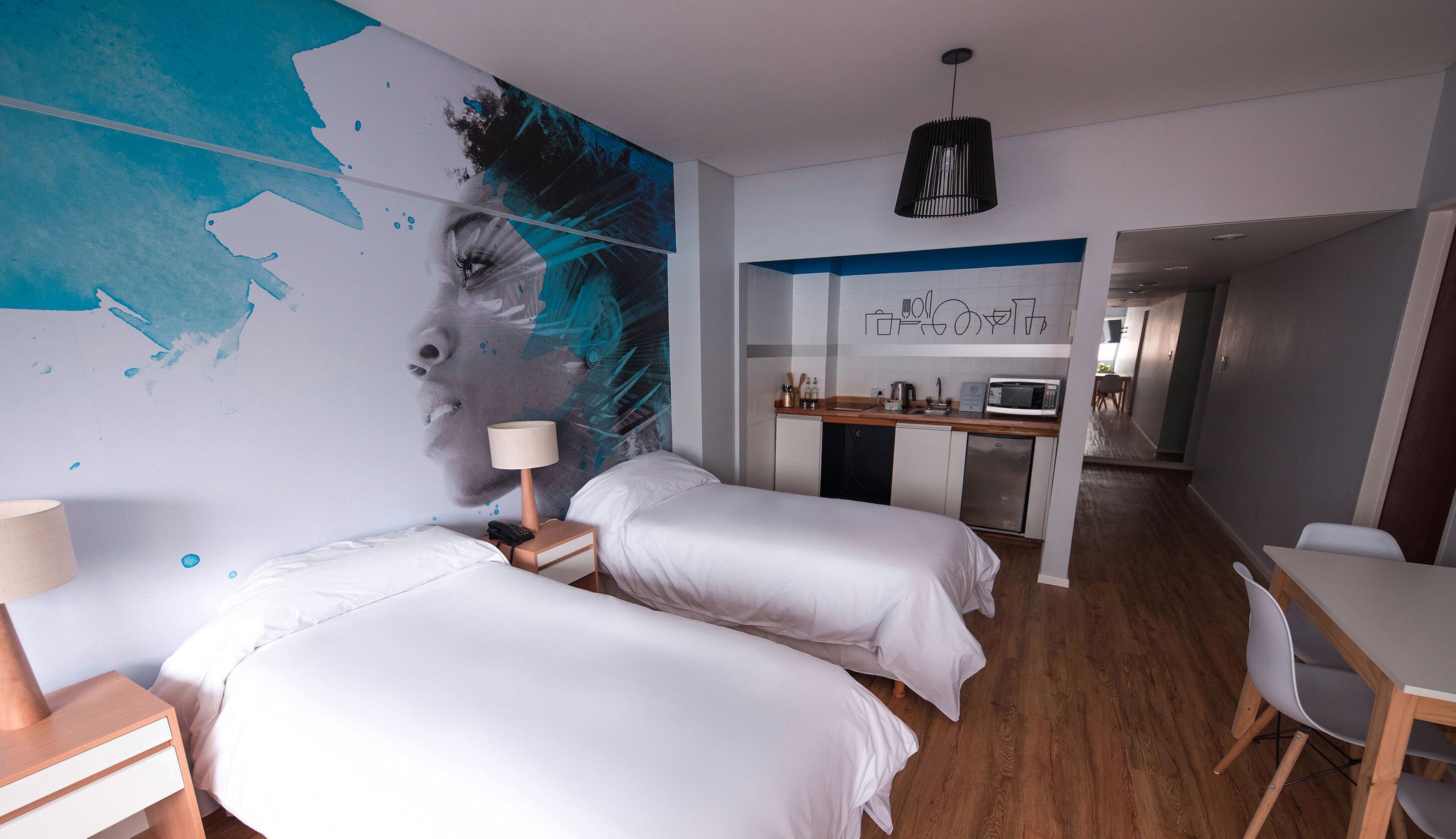 A hotel room with two beds, a desk, a kitchenette, and a bathroom visible in the background.