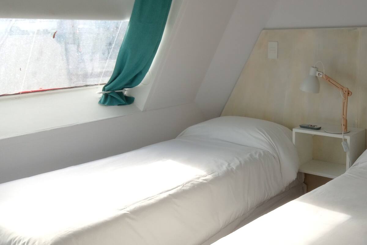 A small, single-room hotel room with two beds, a nightstand, a lamp, and a window with a curtain.
