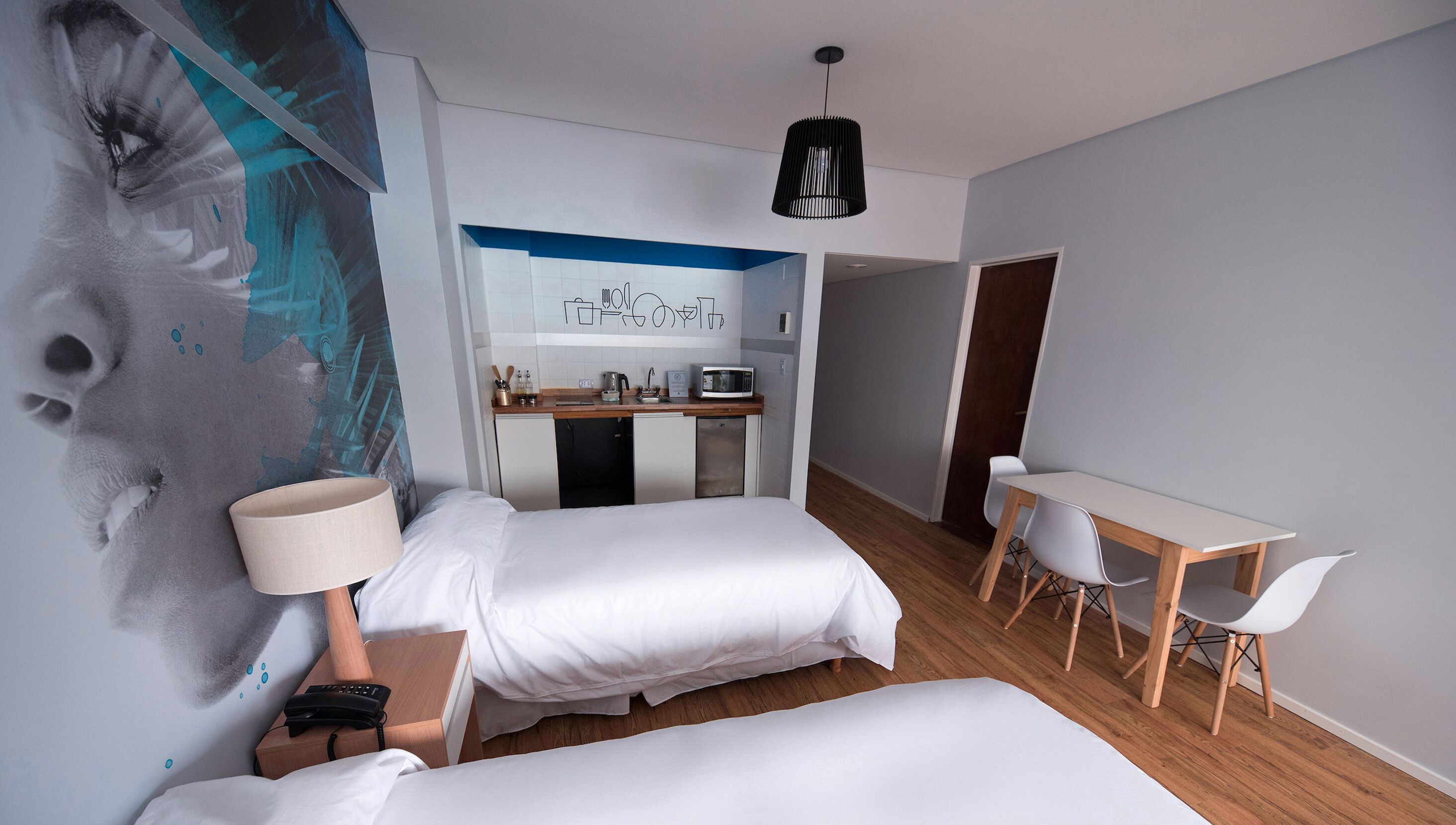 Quarto de hotel com duas camas, uma escrivaninha, uma kitchenette e um mural na parede.