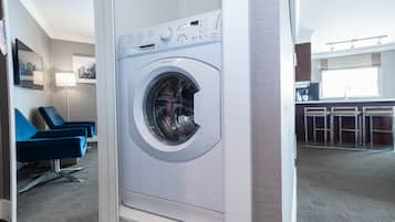 A modern hotel room with a front-loading washing machine, a seating area with blue chairs, and a kitchenette in the background.