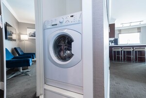 A modern hotel room with a front-loading washing machine, a seating area with blue chairs, and a kitchenette in the background.