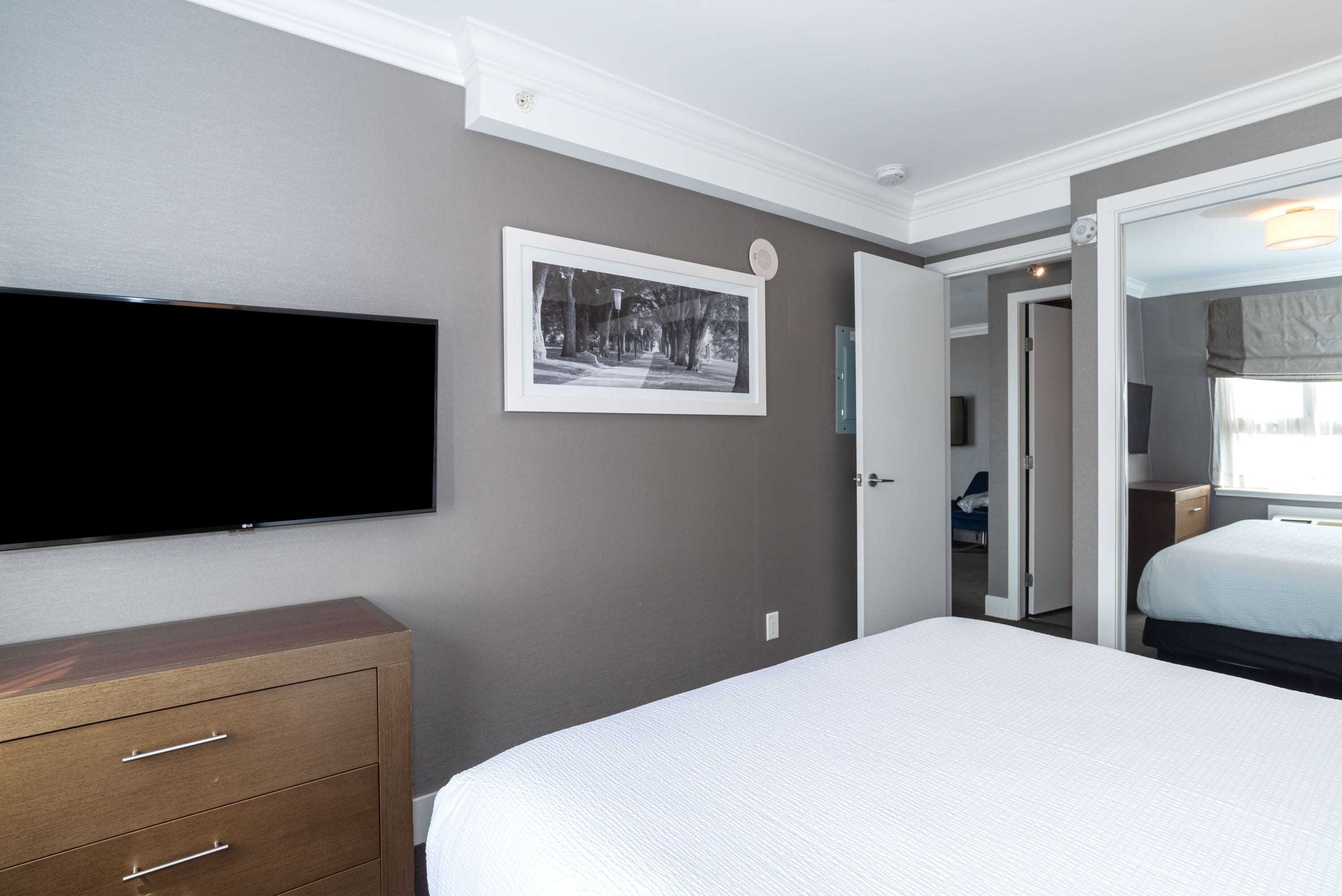 A hotel room with a flat-screen TV mounted on the wall, a wooden dresser, a bed with white bedding, and a framed picture on the wall.