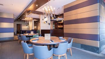 Un espace repas moderne avec une table ronde en bois, entourée de chaises bleues, un lustre décoratif et un mur en pierre agrémenté d’un coin salon en alcôve.