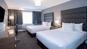 Et hotelværelse med to senge, et skrivebord, en stol, en lampe og et stort vindue med gardiner.
