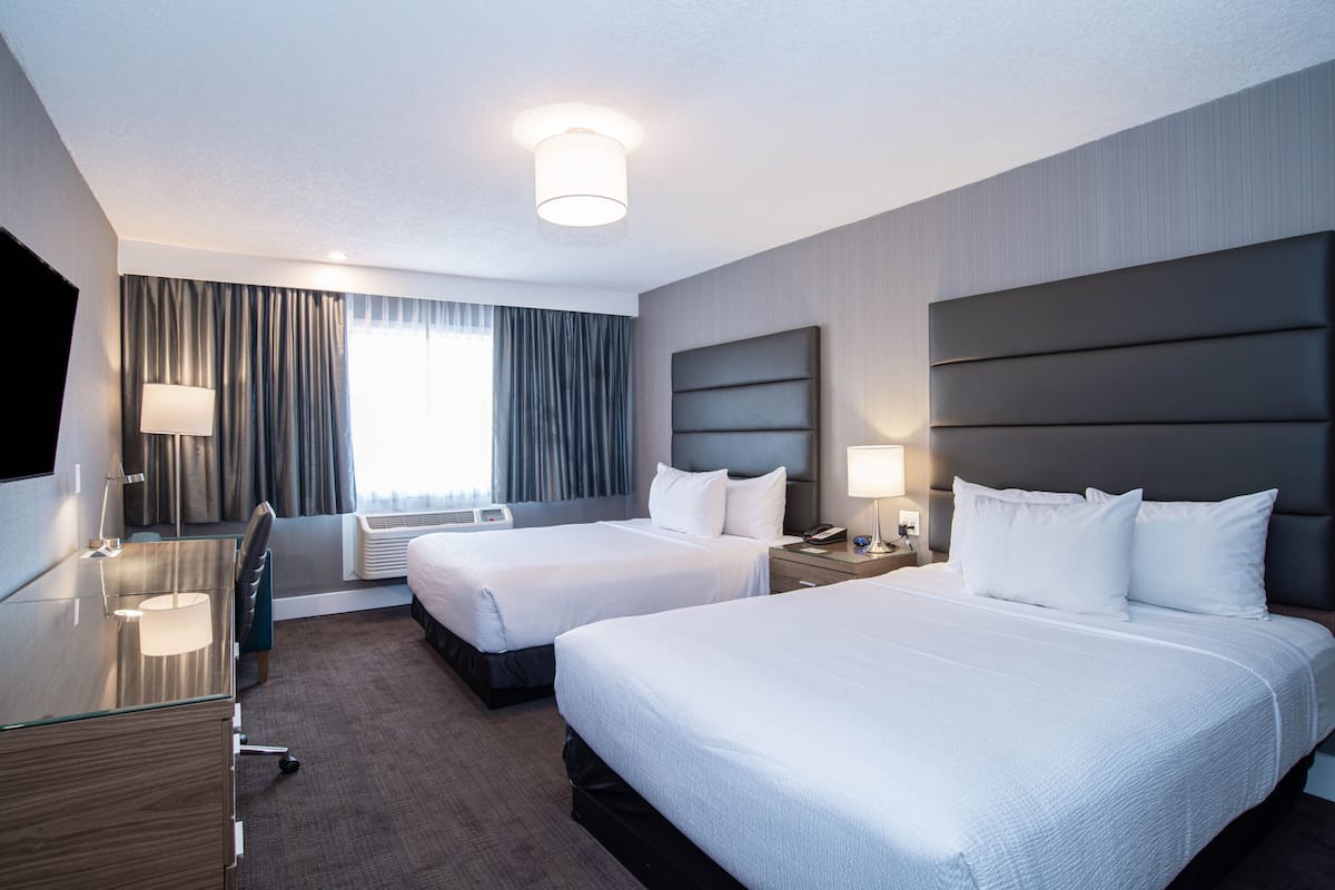 A hotel room with two beds, a desk, a chair, a lamp, and a large window with curtains.