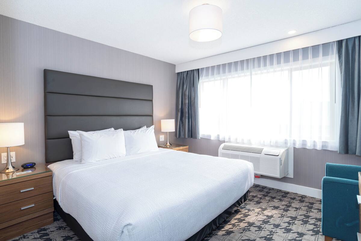 A modern hotel room with a large bed, a bedside table with a lamp, a window with curtains, and a wall-mounted air conditioner.