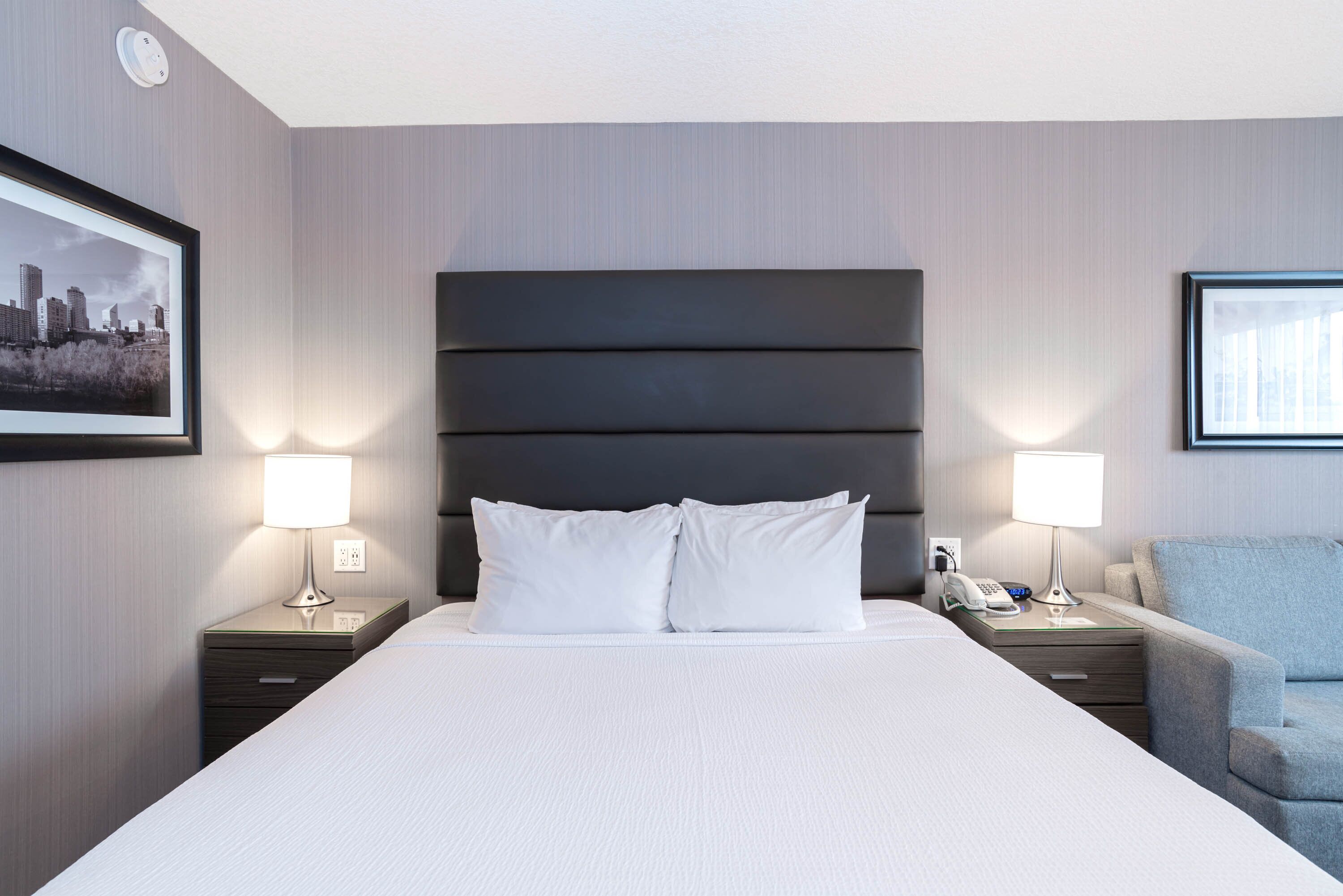 Une chambre d’hôtel avec un grand lit, deux tables de chevet avec des lampes, une chaise et des tableaux encadrés aux murs.