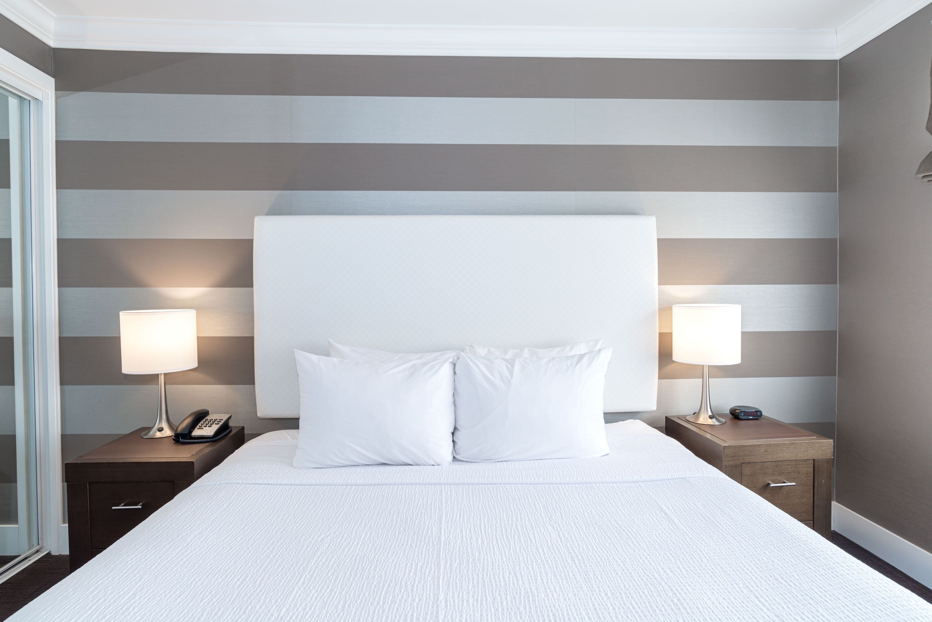 Une chambre d’hôtel avec un grand lit, deux tables de chevet avec des lampes, un téléphone et une petite table de nuit avec une horloge.