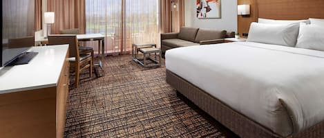 Une chambre d’hôtel avec un grand lit, un bureau avec un ordinateur, une chaise, une petite table et une vue sur l’extérieur grâce à de grandes fenêtres.