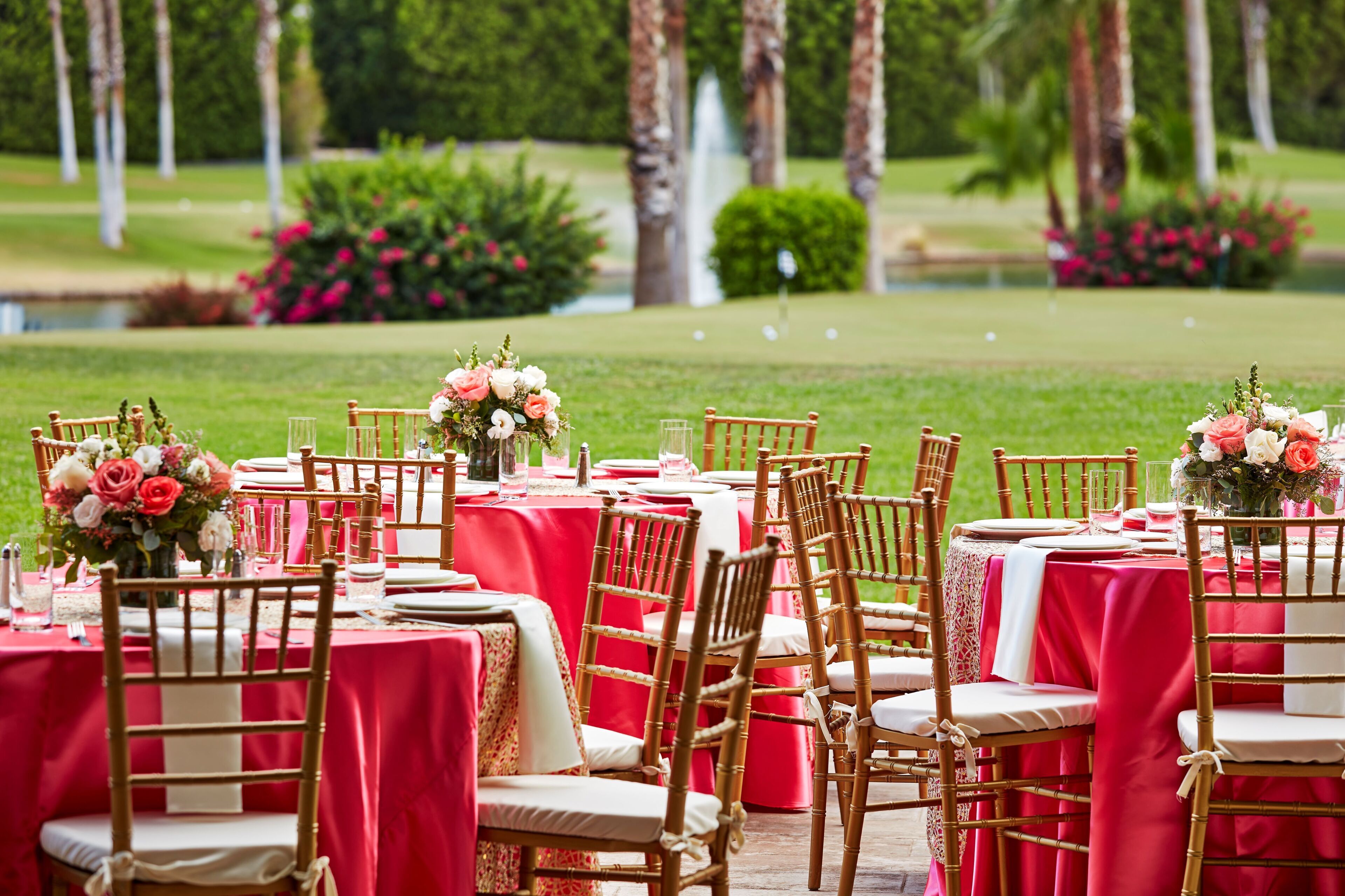 A formal outdoor dining setup with red tablecloths and wooden chairs, arranged in rows with floral centerpieces.