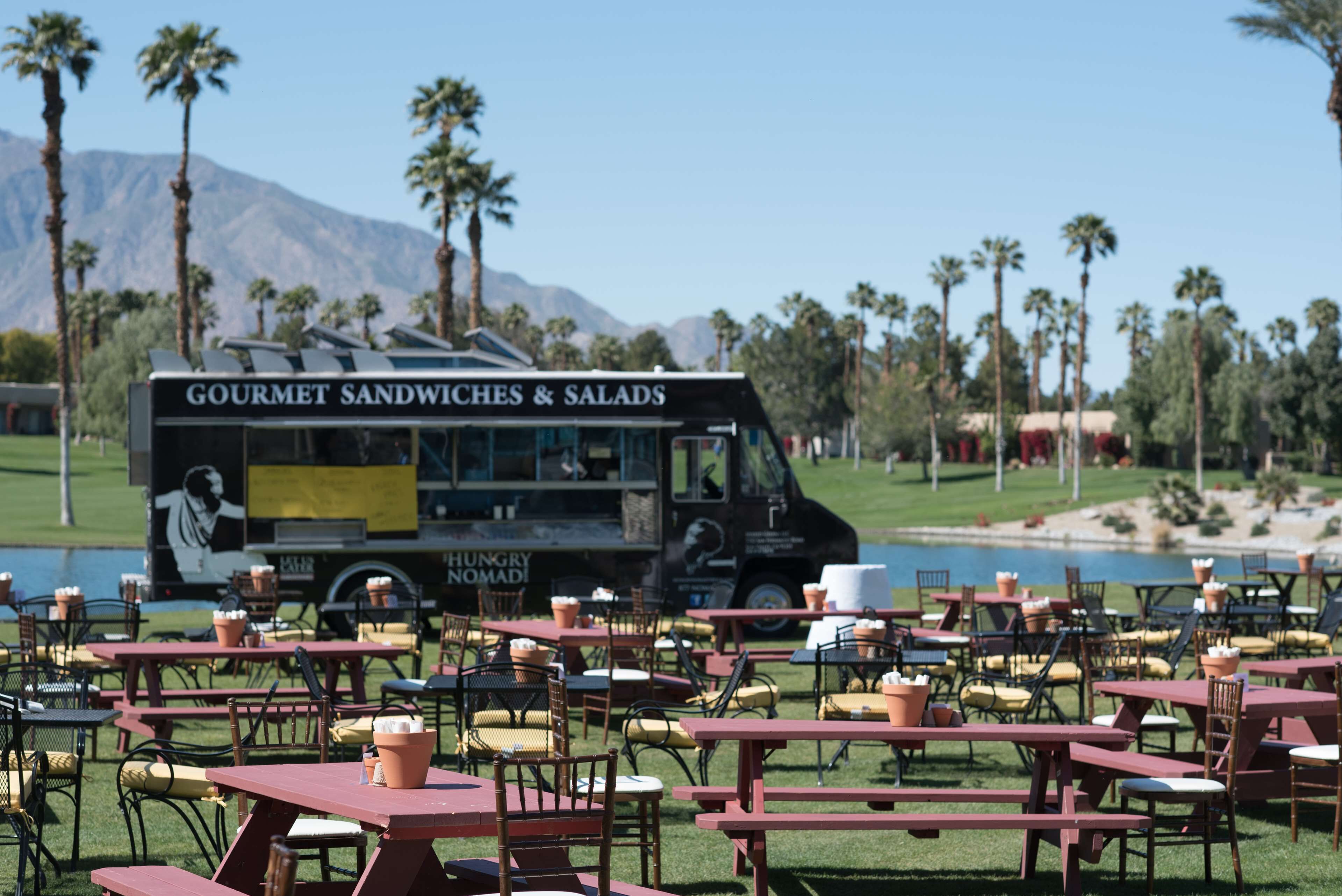 A food truck with a menu for gourmet sandwiches and salads parked in a grassy area with tables and chairs.